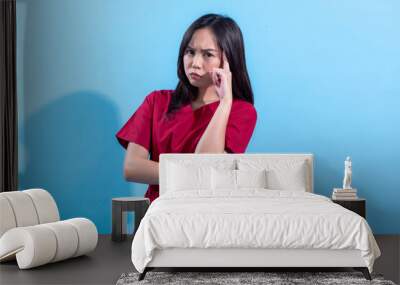 A young Asian woman in a red dress stands with her arms crossed, appearing deep in thought. She touches her temple with one finger, looking pensive, against a light blue background. Wall mural