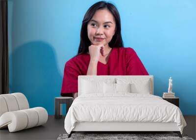 A young Asian woman smiles while thoughtfully resting her hand on her chin. She is wearing a casual red dress and stands against a light blue background, exuding a cheerful and friendly expression Wall mural