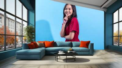 A young Asian woman smiles while thoughtfully resting her hand on her chin. She is wearing a casual red dress and stands against a light blue background, exuding a cheerful and friendly expression Wall mural