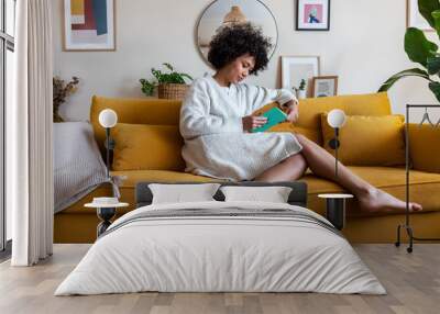 African american woman relaxing at home reading a book sitting on couch. Wall mural