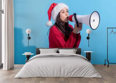 An Asian woman, wearing a Santa hat and a red knitted sweater, is speaking excitedly through a large white megaphone with a red front. She stands against a light blue background. Wall mural