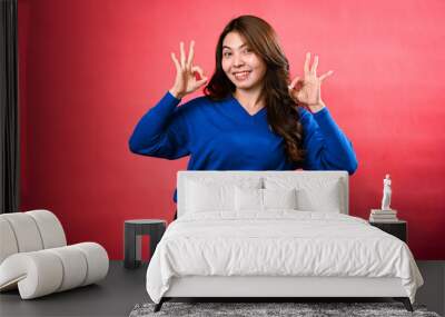 An Asian woman in a blue sweater and black pants poses in front of a red background, making double 