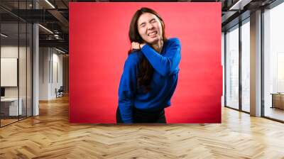 An Asian woman in a blue sweater grimaces in pain, holding her shoulder and back as if experiencing discomfort. She appears to have a muscle ache, standing against a red background Wall mural