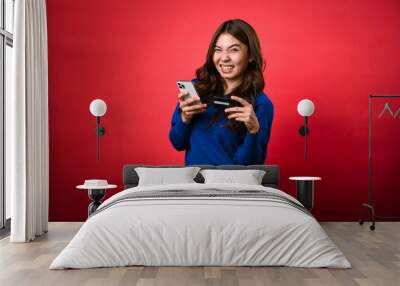 An Asian woman in a blue sweater is happily holding a smartphone and a credit card, looking excited as if making an online purchase. She is standing against a vibrant red background Wall mural