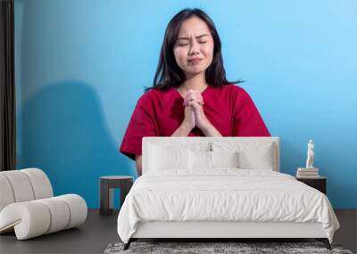 An Asian woman in a red dress stands calmly with her hands clasped together, eyes closed, as if in prayer or gratitude. She appears serene and peaceful against a light blue background Wall mural