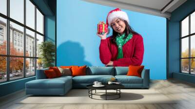 An Asian woman in a red sweater, Santa hat, and green scarf smiles while holding a small, red Christmas gift box with green ribbon and snowflake designs. The background is light blue, Wall mural