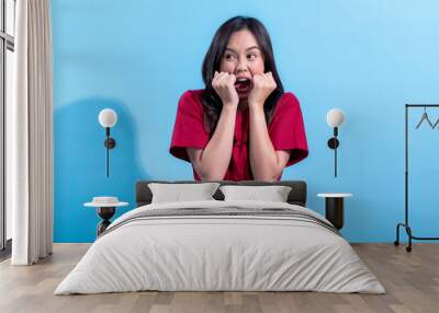 An Asian woman wearing a red dress, expressing fear with her hands clenched near her face and wide eyes, showing a frightened or anxious reaction. She stands against a light blue background Wall mural