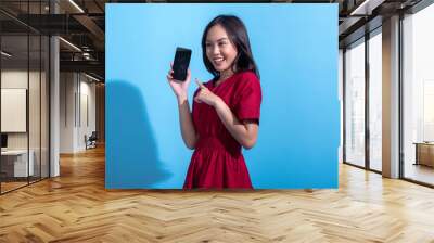 An Asian woman wearing a red dress is smiling and pointing at a black smartphone she is holding in her right hand. The light blue background complements her vibrant red outfit Wall mural