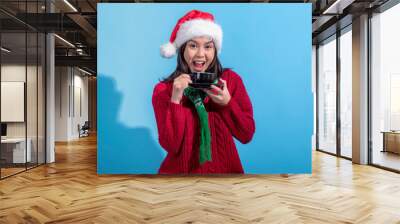 An Asian woman wearing a Santa hat, red sweater, and green scarf with a snowflake pattern is holding a black cup and saucer, smiling excitedly. She poses against a bright light blue background Wall mural