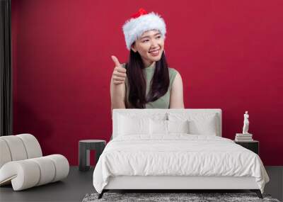 An Asian woman wearing a Santa hat and a sleeveless green dress gives a thumbs-up with a cheerful smile. She stands in front of a solid red background, conveying a festive, positive mood Wall mural