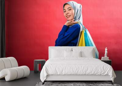 Asian woman wearing a gray hijab and a blue sweater smiles while holding colorful shopping bags in front of a vibrant red background. She looks happy and excited after a successful shopping Wall mural