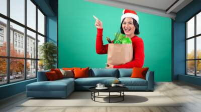 Asian woman wearing a Santa hat and red sweater holds a grocery bag filled with fresh vegetables and points upwards, celebrating the holiday season against a green background Wall mural