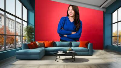 Asian woman with long, wavy brown hair is smiling while standing confidently with her arms crossed. She is dressed in a blue sweater and black pants, posing in front of a red background Wall mural