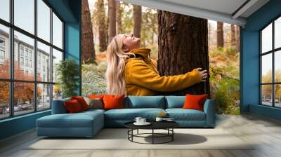 Young blonde woman in yellow coat hugging a tree in the forest and looking up to the tree top Wall mural