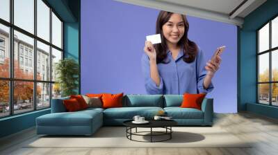 Cheerful Asian woman in blue shirt holding a credit card and smartphone, ready for online shopping, against a purple backdrop Wall mural