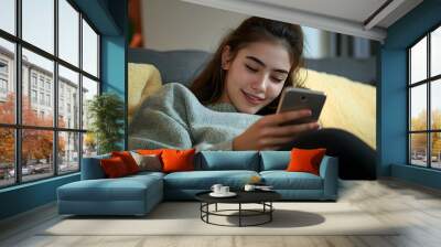 Cheerful young woman reading messages on mobile phone, lying on the sofa at home Wall mural