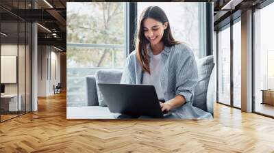 Concentrated millennial girl sit on couch working on laptop browsing internet at home during lazy weekend, focused young woman freelancer busy using computer surfing wireless web shopping online. Wall mural