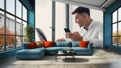 Confident man sitting at desk taking break in work with electronic documents on laptop to make answer telephone call. Smiling young guy freelancer synchronize data between home computer and smartphone Wall mural