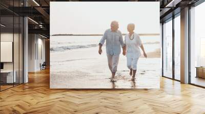 Couple of old mature people walking on the sand together and having fun on the sand of the beach enjoying and living the moment. Two cute seniors in love having fun. Barefoot walking on the water. Wall mural