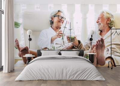 couple of seniors or mature people in a resort spa in their hotel or house clinking with their cocktail or drink - having fun with feet on the table looking between they Wall mural