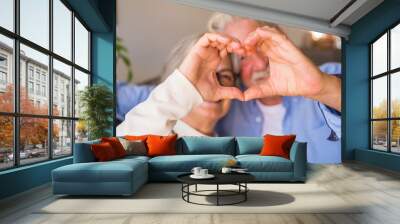 Couple of two old and happy seniors having fun at home on the sofa doing a heart shape with their hands and fingers looking at the camera. . Wall mural