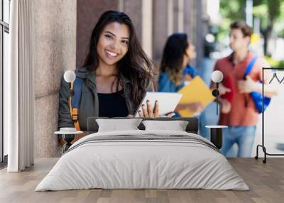 Laughing mexican student with tablet computer and friends in background in front of university building Wall mural