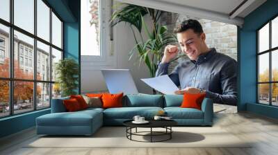 Excited business man student reading postal mail letter overjoyed by great news, happy male winner holding paper bill with loan approval celebrate taxes refund receive salary rise payment sit at desk. Wall mural