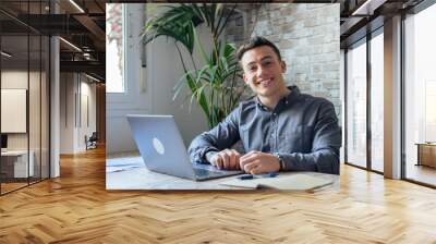 Good-looking millennial office employee in glasses sitting at desk in front of laptop smiling looking at camera. Successful worker, career advance and opportunity, owner of prosperous business concept Wall mural