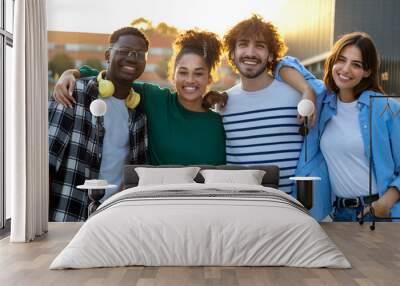 Group of happy and smiling multiracial friends embracing looking at camera standing outdoors during sunset. Wall mural