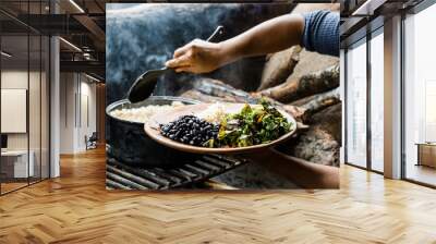 Hands of an unrecognizable woman serving a plate of rice with beans and salad in a traditional kitchen. Wall mural