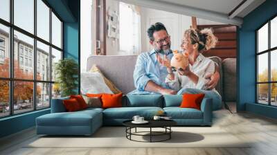 Happy caucasian couple holding piggy bank to save money to make their future dreams come true. Loving man and woman holding piggy bank for savings and discussing while sitting on sofa at home Wall mural