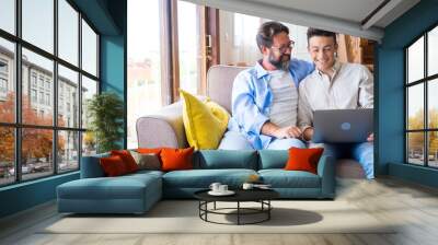 Happy father and son looking at laptop screen while sitting on couch in living room at home. Father with teenage son browsing social media content using laptop in living room Wall mural