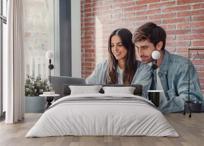 Happy millennial couple sit at table at home browsing web on laptop shopping online together, smiling young husband and wife work on computer at desk pay household bills or taxes in internet banking. Wall mural