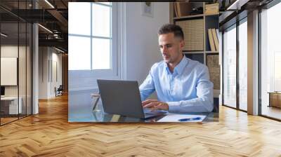 Happy young caucasian millennial businessman working at home at desk with laptop or computer having fun. Young male person in video conference taking care of the business or market.. Wall mural