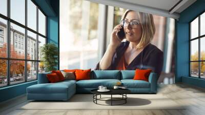 Businesswoman making a mobile phone call to have conversation with management about the company in a modern office. Confident professional blonde female in leadership talking on her smartphone Wall mural