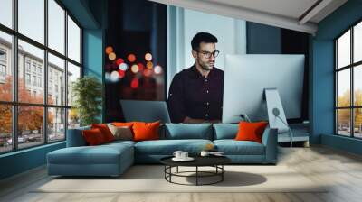 Staying late to get it done. Shot of a young businessman using a computer at his desk during a late night at work. Wall mural