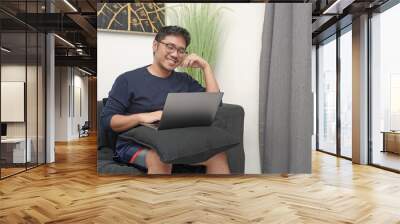 Adult Asian man freelancer sitting in a couch smiling while working using his laptop Wall mural