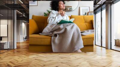 Pensive happy African american woman reading a book at home sitting on sofa relaxing drinking coffee. Vertical image. Wall mural