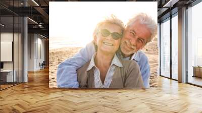 Portrait of couple of mature and old people enjoying summer at the beach looking to the camera smiling and having fun together with the sunset at the background. Two active seniors traveling outdoors. Wall mural