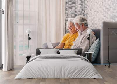 Pretty elderly 70s grey-haired couple resting on couch in living room hold on lap laptop watching movie smiling enjoy free time, older generation and modern wireless technology advanced users concept. Wall mural