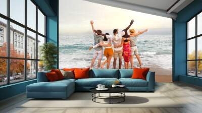 Rear view of multiracial friends embracing together looking at the ocean celebrating with arms up during vacation trip. Wall mural