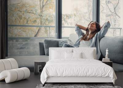 Relaxed serene pretty young woman feel fatigue lounge on comfortable sofa hands behind head rest at home, happy calm lady dream enjoy wellbeing breathing fresh air in cozy home modern living room. Wall mural