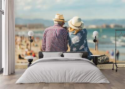 Loving the elderly couple sitting on the wall facing the beach, watching and taking pictures of the landscape on a romantic trip Wall mural