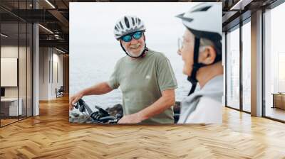 Senior couple riding bikes together in rocky beach enjoying outdoor. Active mature people talking holding mountain bikes. Bicycle lifestyle concept, having fun. Wall mural