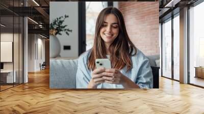 Smiling woman looking at smartphone screen, sitting on couch, enjoying leisure time with mobile device at home, happy young female chatting in social network or surfing internet, shopping online. Wall mural