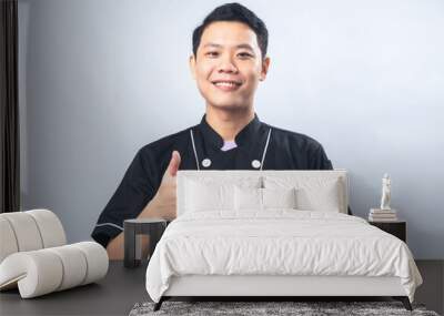 Smiling young man in a black chef uniform, giving a thumbs-up gesture against a plain light gray background. The man exudes confidence and positivity, Wall mural