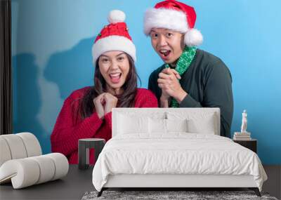 Two cheerful Asian individuals wearing Santa hats and warm outfits, standing with their hands clasped at their chests, celebrating Christmas in front of a light blue background Wall mural