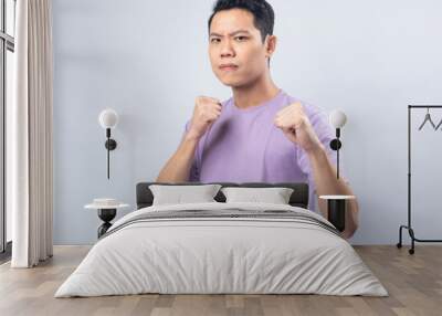 Young Asian man in a lavender t-shirt standing with fists raised, shouting energetically. Studio shot on a plain background emphasizing his excited and victorious expression and casual attire Wall mural