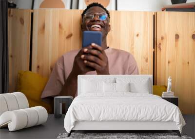 Young black man laughing at home relaxing using mobile phone to watch funny videos. Reading text message. Wall mural