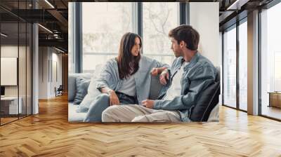 Young happy couple having fun talking laughing relaxing at home on couch, boyfriend embracing girlfriend telling funny joke sitting on sofa, humor in relationships, enjoying weekend together. Wall mural